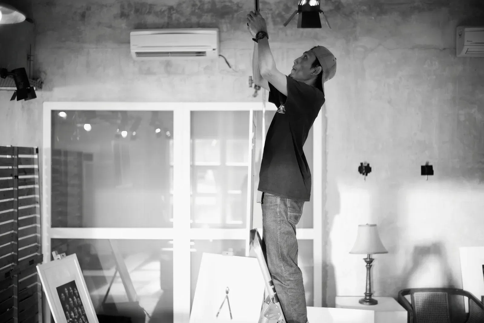 An electrician standing on a ladder and working on an overhead light fixture indoors